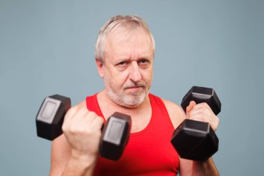 Senior man adds humor to fitness A caucasian male pensioner stands in the gym, lifting dumbbells while making a funny face. The blue background provides a unique touch to this amusing