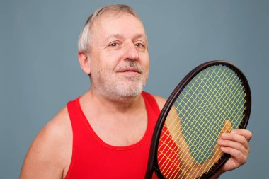 Elderly Athlete in Studio with Tennis Racket This image captures a senior man who is defying his age with his active lifestyle. The man is seen holding a tennis racket and is dressed in a red tank top