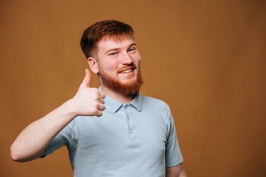 A caucasian young man gives a thumbs up and looks straight at the camera with a smile on his face in a studio setting. An adolescent male with red hair looks satisfied and happy while flashing