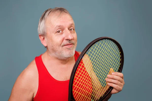 Senior Men Enjoying Tennis A pensioner who is still active in sports, is captured in this image. The man is seen holding a tennis racket, dressed in a red tank top and is set against a blue background