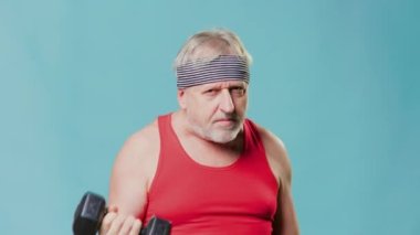 Senior man adds humor to fitness A caucasian male pensioner stands in the gym, lifting dumbbells while making a funny face. The blue background provides a unique touch to this amusing
