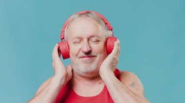 An aged senior citizen takes a break from his daily routine, enjoying a good laugh while using his earphones to listen to his favorite tunes. Blue studio background emphasizes his lighthearted