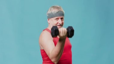 Pensioner in the gym proves age is just a number, studio shot an elderly man with a sporty and energetic demeanor. Holding dumbbells and looking directly at the camera with a humorous expression,