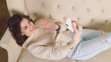 Memnuniyetle kanepede oturan bir kadın, akıllı telefonunu kullanarak iş arkadaşlarıyla sanal bir toplantı yapıyor ve büyük bir gülümseme sergiliyor. Evde Gülümseyen Gençler Uzaklık Video Çağrısı
