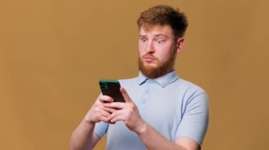 Stüdyodaki genç kızıl saçlı bir adam yüzünde korkmuş bir ifadeyle akıllı telefonuna bakıyor ve şok olmuş bir ifadeyle akıllı telefonuna bakıyor.