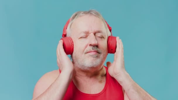 高齢者の男性は 彼のイヤフォンを介して音楽を聞いている 驚きと何かHを聞いて驚いている スタジオの青い背景は彼の表現とは対照的である — ストック動画