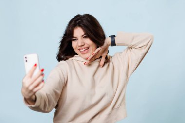 A young adolescent girl is seen in the photo, sitting on a blue background in a studio. She is a blogger and vlogger, and is captured in a moment of happy conversation and greeting,