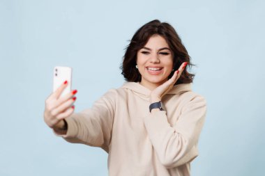 A young adolescent girl is seen in the photo, sitting on a blue background in a studio. She is a blogger and vlogger, and is captured in a moment of happy conversation and greeting,