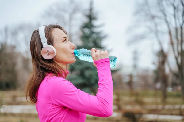 An athletic woman running on a trail in taking a moment to drink water from her bottle. A real people jogger, a female runner in her 20s, drinking water from her bottle