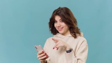 Gülümseyen esmer bir genç kızın portresi mavi arka planda izole bir şekilde kameraya bakıyor, akıllı telefon gösteriyor, işaret ediyor. Aramalar için yeni uygun tarifeler. Boşluğu kopyala