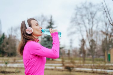 A real people jogger, a woman in her 30s, running and drinking water while maintaining her healthy lifestyle. A female athlete taking a break from her outdoor running session, drinking water