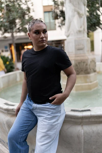 Non-binary person in makeup looks away while sitting near an outdoor water fountain. LGBTIQ concept.
