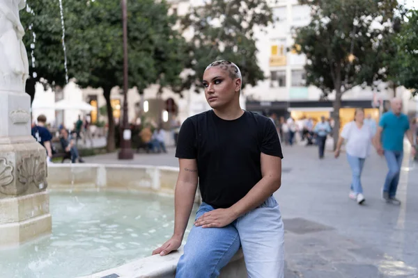 A non-binary person sitting near an outdoor water fountain. LGBTIQ concept.