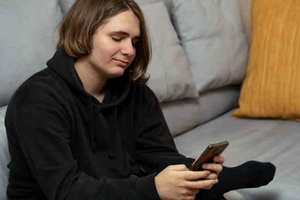 stock image Young man with cheeky smile using a smartphone. Young man is sitting smiling cheekily while looking at the smartphone he is holding in his hands.