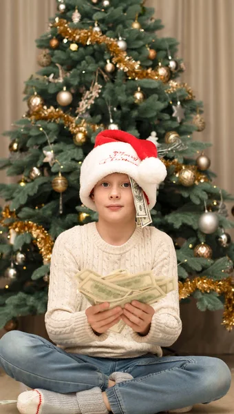 Christmas concept. Black Friday. A handsome caucasian boy in a red Santa Claus hat holds a lot of paper money in his hands against the background of a chic green Christmas tree decorated with