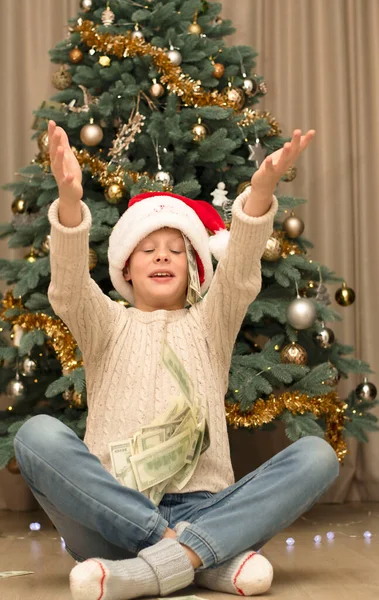 Christmas concept. Black Friday. A handsome caucasian boy in a red Santa Claus hat catches flying paper money with his hands against the background of a chic green Christmas tree decorated with New