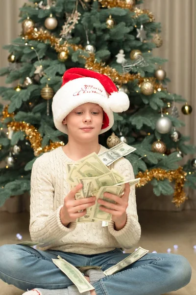 Christmas concept. Black Friday. A handsome caucasian boy in a red Santa Claus hat holds a lot of paper money in his hands against the background of a chic green Christmas tree decorated with