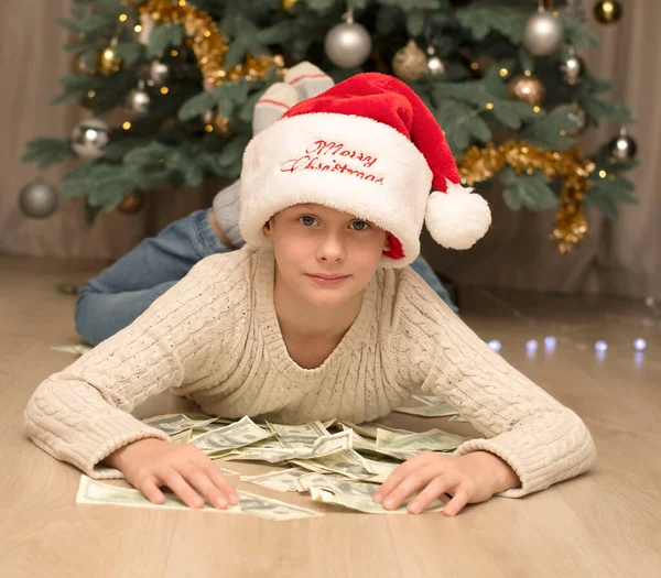 Christmas concept. Black Friday. A handsome boy in a red santa claus hat lies on the floor with a huge pile of paper money against the background of a smartly decorated Christmas tree decorated with