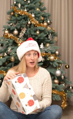 - İnsanlar. Tatil günü. Noel konsepti. Noel Baba şapkalı güzel beyaz kadın bir Noel ağacının arka planında duygusal olarak sürpriz bir hediye kutusu açıyor..