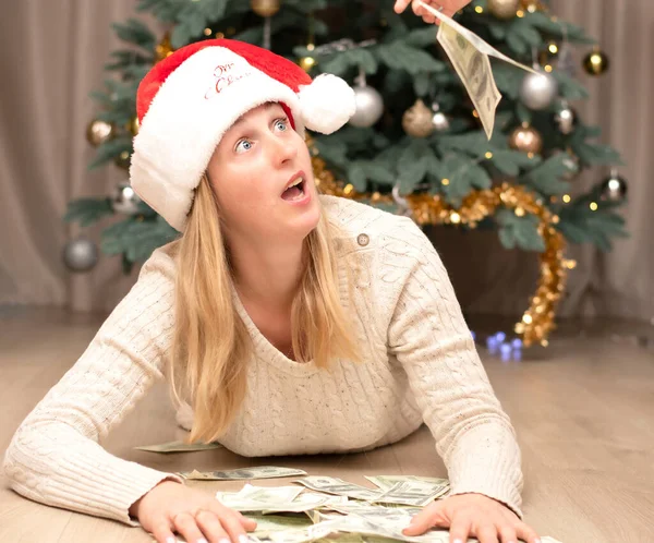 Caucasian Beautiful girl in a santa claus hat lies on the floor with a pile of paper money on the background of a Christmas tree. Christmas concept. Black Friday concept