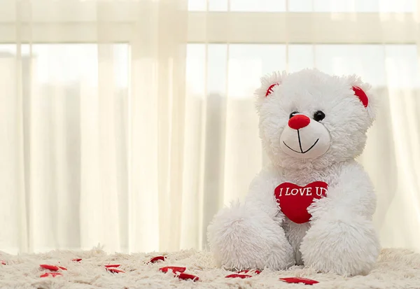 Valentine\'s day concept. A cute white teddy bear sits on a window rug with a bright red heart. Close-up. soft focus