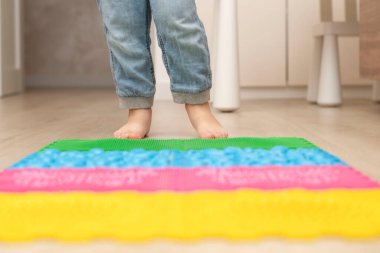 Health concept. A little boy stands with bare feet on a multi-colored orthopedic massage mat in a home interior. Close-up of childrens heels. Back view.