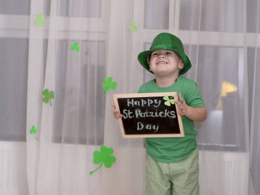 St. Patricks Day concept. A small cheerful caucasian boy in a shiny green top hat and green clothes holds a black wooden board with an inscription in chalk the holiday of St. Patrick on the