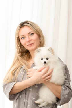 İnsanlar ve hayvanlar. Güzel ve mutlu bir Kafkas kızı, uzun saçlı bir sarışın, kollarında, doğal kumaştan yapılmış gri ev yapımı pijamalarıyla Japon Spitz cinsinden şirin beyaz bir köpek tutuyor.