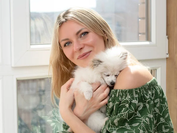People and animals. A beautiful girl, a blonde in green home clothes, holds a white small dog of the Spitz breed and gently hugs her against the background of the window. Close-up. Clncept.