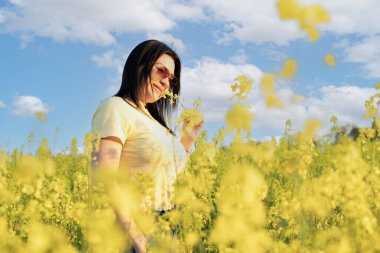 Güneş gözlüklü güzel bir kız mavi gökyüzüne karşı sarı çiçekli bir tarlada duruyor, gülümsüyor ve kolza çiçeği kokuyor. Barış kavramı. Yumuşak odaklanma. Boşluğu kopyala.