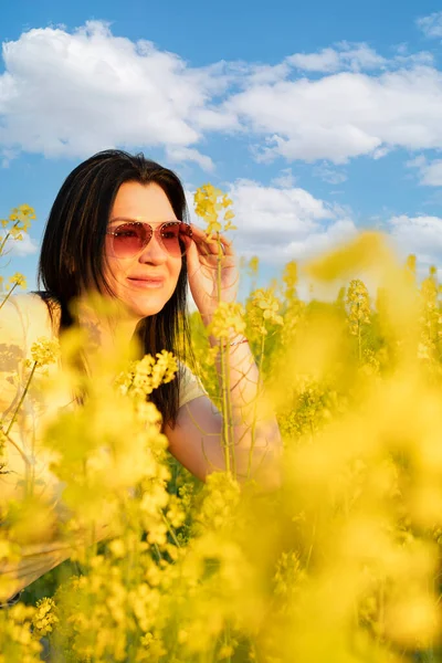 A beautiful girl in sunglasses stands in a field of yellow flowers against a blue sky, smiles happily and inhales the fresh spring and clean air. Peace concept. The color of the flag of Ukraine. No