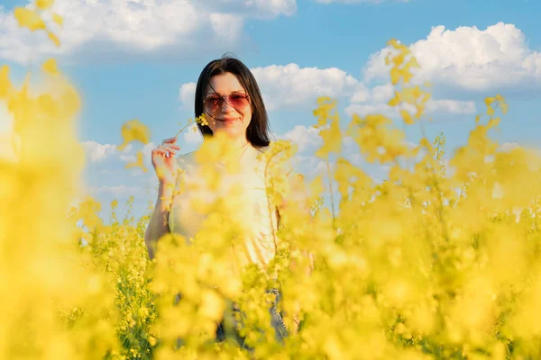 A beautiful girl in sunglasses stands in a field of yellow flowers against a blue sky, smiles happily and inhales the fresh spring and clean air. Peace concept. The color of the flag of Ukraine. No