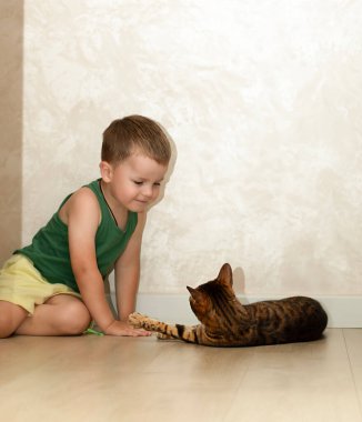 4 yaşında güzel bir çocuk, kırmızı bir leopar bengal kedisiyle evin içinde oynarken eğleniyor. Evcil kedi yerde yatar ve pençesiyle bebekle oynar..