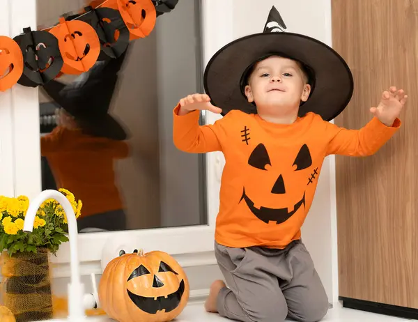 Halloween concept. A small, cheerful, handsome boy in a wizard\'s hat and an orange sweater sits on a table in the kitchen against the background of a pumpkin, a ghost and a bouquet of yellow flowers. Festive interior.