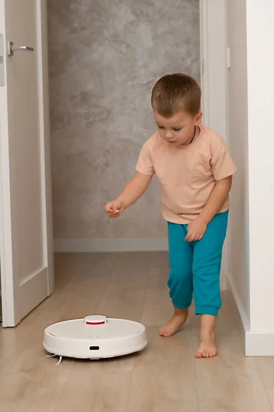 Cleaning concept. A little funny Caucasian boy, 4 years old, throws torn paper in small pieces onto the floor for cleaning and suction by a white robot vacuum cleaner. Plays happily in the home
