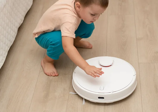 A little cheerful boy in home clothes, throws torn paper into small pieces on the floor for cleaning and suction by a white robot vacuum cleaner. Enjoys playing in the home interior.