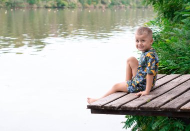 Balık tutma konsepti. Duygusal yüz ifadeleri olan küçük yakışıklı bir çocuk, 4 yaşında, nehir kenarındaki ahşap bir köprüde oturuyor, yeşil bitkilerin arka planına yaslanıyor ve temiz hava soluyor. Rahat ol. Yer