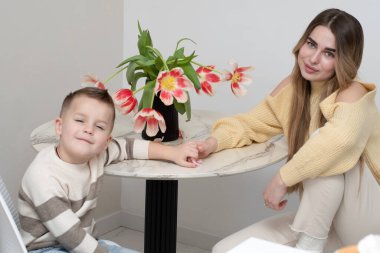 Aile kavramı. 19 yaşında güzel bir kız ve 5 yaşında bir oğlan beyaz bir arka planda kırmızı lale buketiyle yuvarlak masada büyüleyici bir şekilde oturuyorlar. Erkek ve kız kardeş bağı. Sarılmalar ve aile bağlantısı.