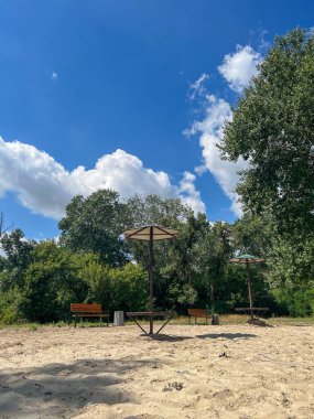 Boş banklar ve piknik masaları şemsiyelerin altında kumlu bir sahilde, mavi gökyüzünün altında yeşil ağaçların altında biraz bulutlu.