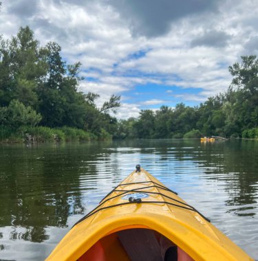 Sarı kano, yemyeşil ağaçların ve bulutlu gökyüzünün ortasında huzurlu bir kano deneyimi sunarak huzurlu bir nehirde geziniyor.