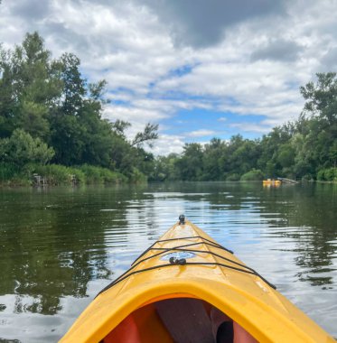 Sarı kano, yemyeşil yemyeşil, bulutlu bir gökyüzünün altında sakin bir kano deneyimi sunuyor.