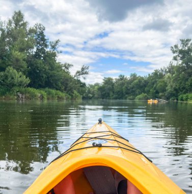 Kanocu bulutlu bir gökyüzünün altında sakin bir nehirde kürek çekiyor, sarı bir kanoyla huzurlu bir su yolculuğunun tadını çıkarıyor.