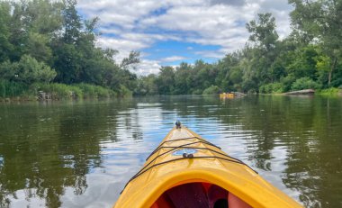 Sarı kano güneşli yaz gününde sakin bir nehirde süzülüyor her iki tarafında yemyeşil ağaçlar var.