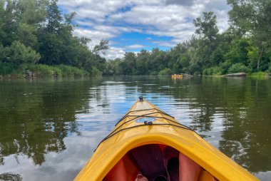 Sakin bir nehirde kürek çeken kanocu bulutlu bir gökyüzünün altındaki yemyeşil ağaçların huzurlu manzarasının tadını çıkarıyor.