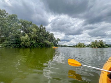 Sarı kano, yeşil ağaçlar ve adalarla çevrili sakin nehir sularında dramatik bulutlu bir gökyüzünün altında yüzüyor.