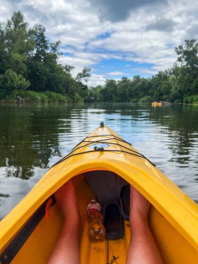 Sarı kanoyla nehirde kürek çeken kanocu, bereketli ağaçlar ve bulutlu gökyüzü ile sakin doğa manzarasının tadını çıkarıyor.