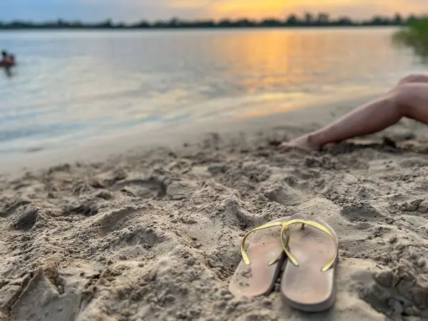 Gün batımında kumlu sahilde terlikleriyle dinlenen bir kadın.