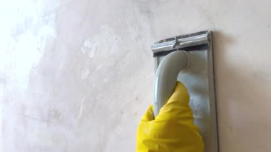 İnşaat işçisi kumlu gri bir duvarı zımpara makinesiyle zımparalıyor ve sarı eldivenler giyiyor.