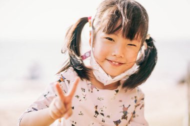 Asyalı kızın portresi açık havada zafer işareti kaldırdı. Pan-Asyalı küçük tatlı kız gülümseyip kameraya bakıyor..