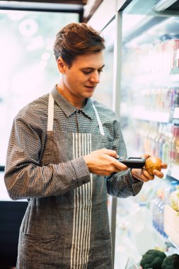 Marketteki taze meyve dükkanında gizli hayat tarzı genç bir adam raftaki ürünlerin kalitesini kontrol ediyor. Maskesiz genç bir erkek, mini sanatlarda çalışıyor..